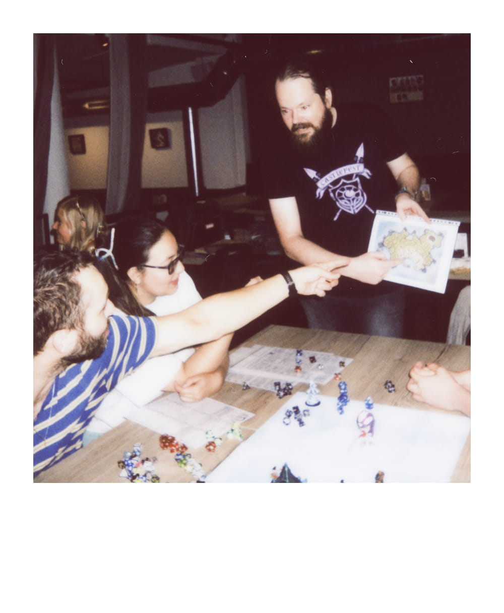Game Master shows a fantasy map as a player points across a table covered with dice and miniatures.